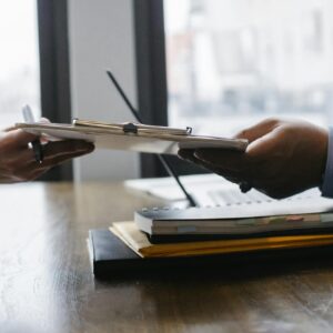 Crop anonymous ethnic woman passing clipboard to office worker with laptop during job interview