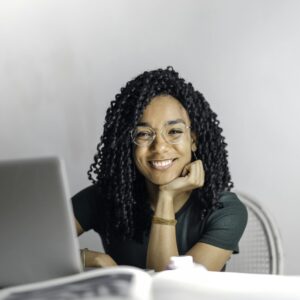 Joyful businesswoman with curly hair smiling at camera while using laptop indoors.